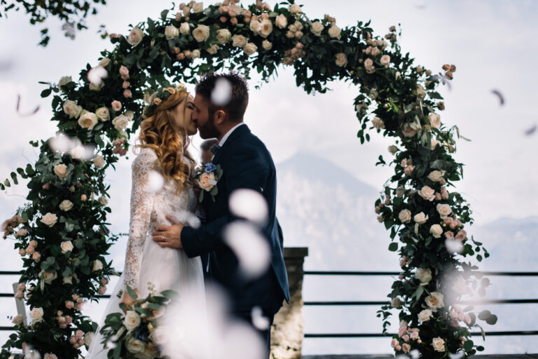 Videograf Hochzeit – romantischer Kuss des Brautpaars unter Blumenbogen